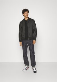 Giacca bomber nera con una texture liscia, chiusura con zip, polsini a costine, abbinata a jeans scuri e sneakers nere, bianche e rosse.