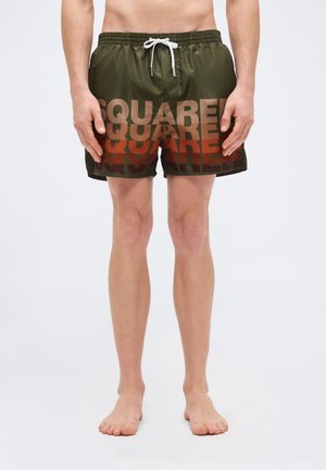 Homme portant un short de bain vert olive avec le texte répétitif "SQUARE" en dégradé du brun au rouge, debout pieds nus sur un fond blanc.