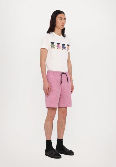Witte T-shirt met een multicolore grafische print, gecombineerd met roze patroon shorts en zwarte sandalen met dikke banden. Zwarte trekkoord aan de shorts.