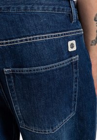 Jean en denim bleu foncé présentant une poche arrière avec une couture blanche contrastante et une petite étiquette de marque. Texture lisse avec une coupe classique.