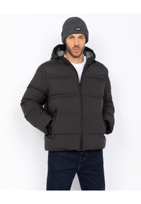 Mand med en sort polstret jakke med hætte, blå jeans, en grå strikket hue og hvid skjorte, der står med hænderne i jakke-lommerne.