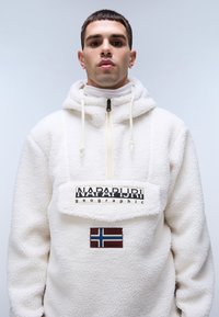 Uomo che indossa una felpa bianca con cappuccio con la scritta "NAPAPIJRI geographic" e una toppa con la bandiera nordica sulla parte anteriore, su uno sfondo chiaro e semplice.