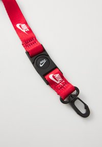 Červený nylonový popruh s černou plastovou sponou s kruhovým designem a černou kovovou sponou na konci. Na popruhu je vytištěno logo Nike v bílé barvě.