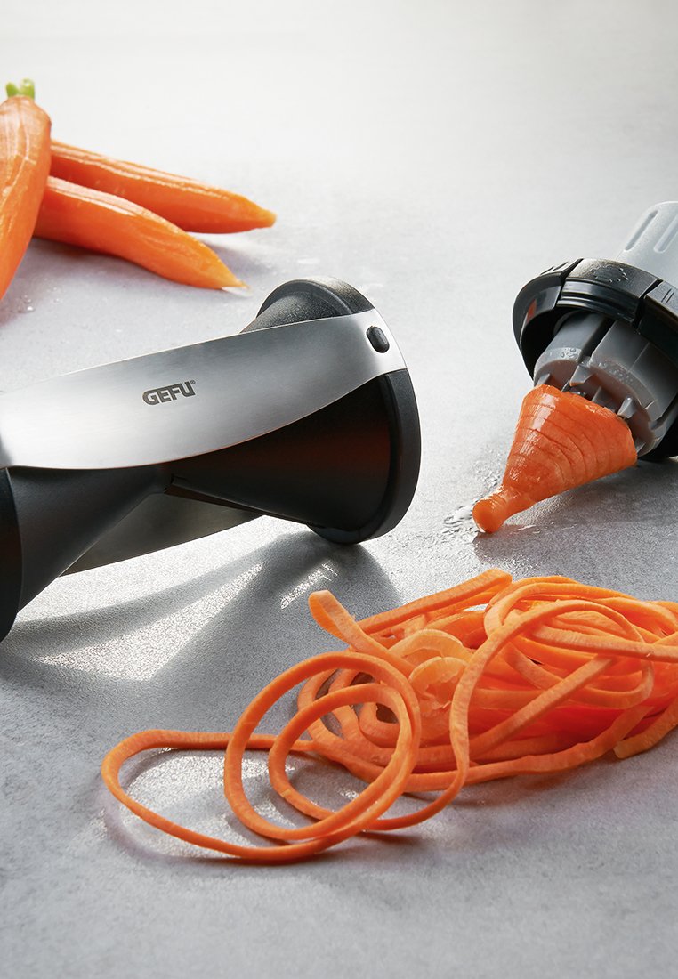 GEFU Spiralschneider mit einer Klinge aus Edelstahl und einem schwarzen ergonomischen Griff. Gewundene orange Karottenstreifen und ganze Karotten sind in der Nähe.