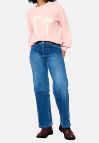 Pull rose clair avec motif de palmier blanc et décorations, associé à un jean bleu taille haute à jambe droite et des bottes marron.