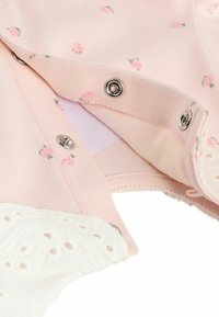 Helles rosa Kleidungsstück mit kleinen Blumenmustern, ausgestattet mit Metall-Druckknöpfen. Der untere Teil hat einen weißen Spitzenbesatz mit dekorativen Löchern.