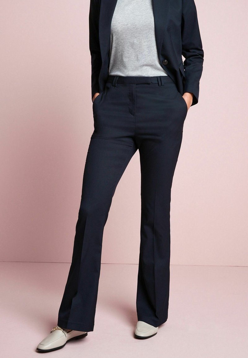 Femme portant un pantalon de costume bleu marine, un blazer assorti, un t-shirt gris et des mocassins blancs, debout les mains dans les poches devant un fond rose.