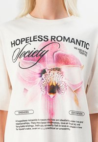 Krémové bavlnené tričko s veľkým grafickým motívom ružovej orchidey a čiernym textom. Detaily obsahujú "NÁDEJNÝ ROMANTIK SOCIETY" nad a definície nižšie.