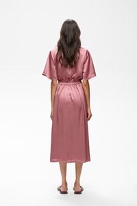 Femme aux longs cheveux foncés portant une robe midi en satin rose à manches courtes, debout de dos sur fond blanc.