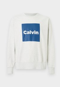 Svetlošedý teplák s modrým obdĺžnikovým logom, na ktorom je slovo "Calvin" v bielej farbe. Mäkká tkanina, rebrované manžety a lem. Neformálny dizajn.