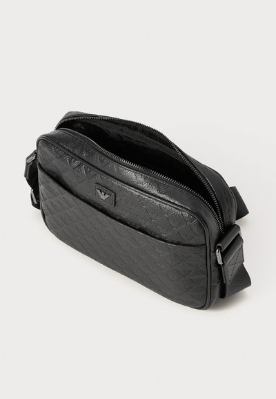 Geantă crossbody texturată neagră, cu buzunar frontal cu fermoar, curea ajustabilă și model logo în relief pe fundal alb.