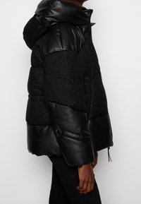 Černá péřová bunda s kombinací texturované tkaniny a hladkých kožených rukávů, s kapucí a nastavitelním stahováním na dolním lemu.