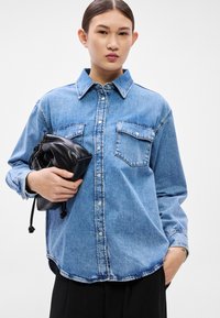 Svetlomodrá džínsová košeľa s dvoma náprsnými vreckami, zapínaním na gombíky a mierne voľným strihom. Drží malú čiernu koženú kapsičku.
