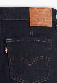 Jeans in denim scuro con etichetta in pelle impressa con "Levi Strauss & Co." e cuciture in rame. Presenta una sola tasca posteriore con accento in tessuto rosso.