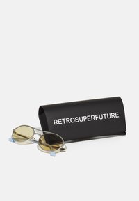 Occhiali da sole rotondi in argento con lenti gialle, con punte azzurre chiare, accompagnati da una custodia protettiva nera con testo bianco.