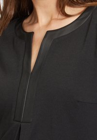 Gros plan sur une femme portant un haut noir avec un décolleté en V souligné par une bordure en cuir noir.