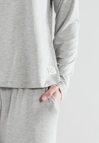 Grijze lange mouwtop met een losse pasvorm, gemaakt van zachte stof. Heeft een zak en een klein merklabel aan de zijkant. Gecombineerd met bijpassende grijze broek.