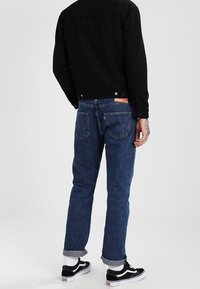 Giacca di denim nera, jeans blu con risvolti, vista posteriore. Cuciture evidenti, tasche e marchio sulla giacca. Scarpe Vans visibili.