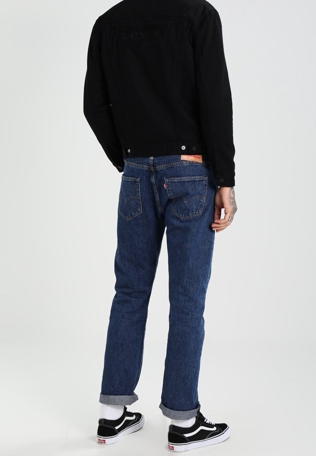 Giacca di denim nera, jeans blu con risvolti, vista posteriore. Cuciture evidenti, tasche e marchio sulla giacca. Scarpe Vans visibili.