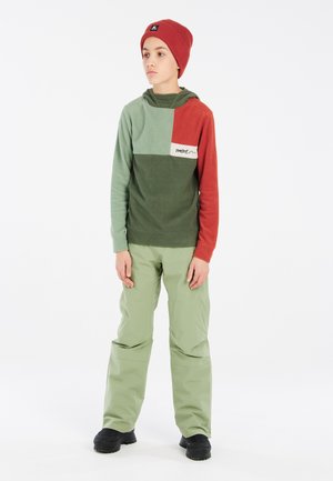 Sweatshirt à capuche vert et rouge, avec un design texturé et un accent logo. Associé à un pantalon vert clair et des chaussures noires.