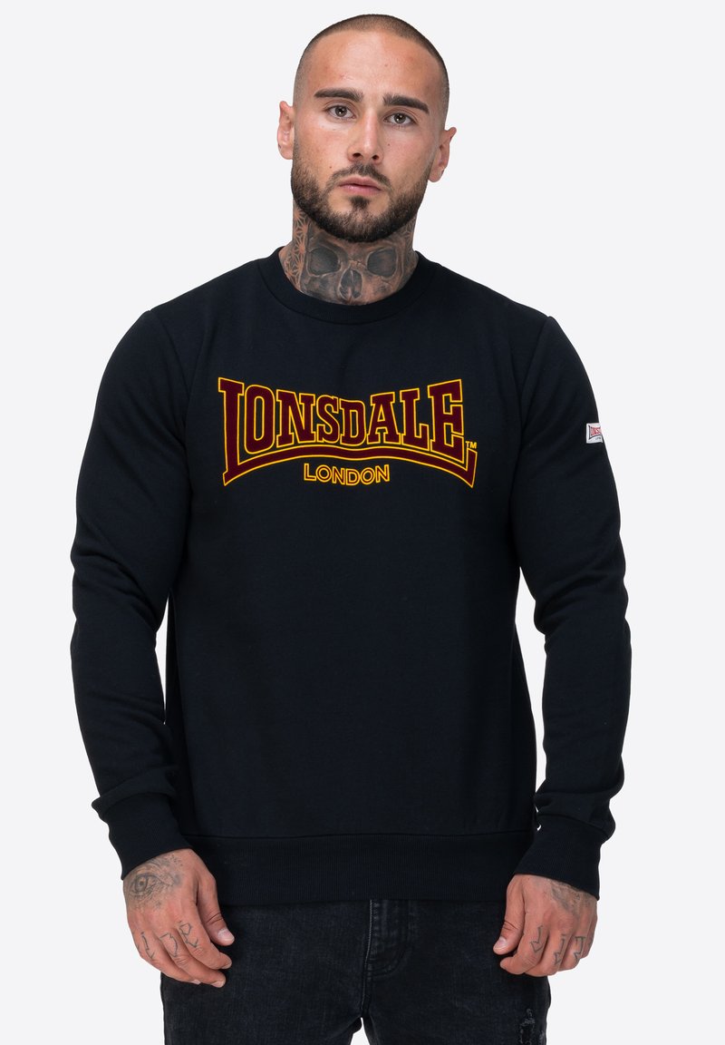 Sudadera negra con un prominente logo amarillo y rojo de "Lonsdale London", puños y dobladillo acanalados, y una textura suave.