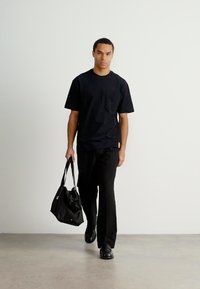 Μαύρη βαμβακερή μπλούζα T-shirt με τσέπη, σε συνδυασμό με μαύρο παντελόνι. Το μοντέλο κρατάει μια μαύρη τσάντα, στέκεται σε ανοιχτό γκρι δάπεδο μπροστά από έναν λευκό τοίχο.