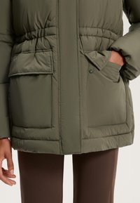 Olíva zöld párnázott dzseki, megkötött derékkal, két elülső zsebbel és látható varrással. Puha tapintású és középhosszú fazon.