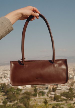 Hånd, der holder en brun lædertaske med langt håndtag, byens skyline i den slørede baggrund under klar himmel.