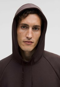 Brauner Hoodie mit einer Kordelzugkapuze, der eine glatte Textur und Raglanärmel aufweist. Der Stoff wirkt weich und leicht dehnbar.