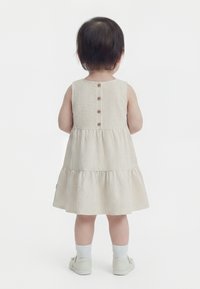 Småbarn, set bagfra, iført en beige, stribet ærmeløs kjole med knapper ned ad ryggen, hvide sokker og beige sko.