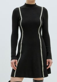 Schwarzes geripptes Strickkleid mit langen Ärmeln, hohem Kragen und weißen Zickzackstichen entlang der Seiten, ausgestellt am Saum.