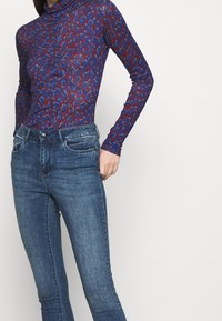 Top de manga larga en estampado de leopardo azul y rojo, hecho de tela elástica. Combinado con jeans skinny de mezclilla oscura que cuentan con bolsillos delanteros y cierre de botón.