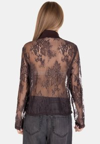 Donna con capelli lunghi che indossa una blusa in pizzo nero trasparente con motivi floreali e jeans neri sbiaditi, vista di spalle.