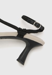 Schwarze und beige High-Heeled Sandale mit einem geflochtenen schwarzen Riemen und einer verstellbaren Schnalle. Glattes Lederoberteil mit einem eleganten Fersendesign.