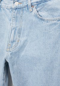 Gros plan sur un jean en denim bleu clair montrant la poche avant, le bouton en métal, la passants de ceinture et les détails de couture visibles.