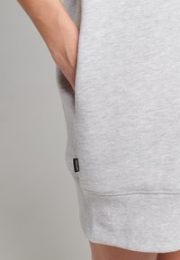 Hellgraues Sweatshirtkleid mit geripptem Saum, seitlichen Taschen und einem kleinen schwarzen Etikett. Der Stoff zeigt eine dezente Textur.