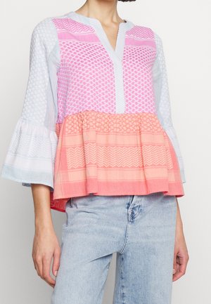 Kvinde iført en løs, mønstret bluse med sektioner i pink, koral og lyseblå samt klokkeærmer, kombineret med lyseblå jeans.