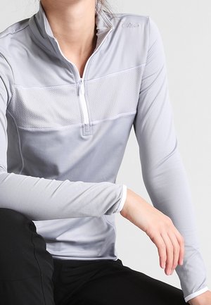 Person iført en lysegrå langærmet sportstop med kvart lynlås og tekstureret brystpanel, siddende med den ene arm hvilende på et knæ.