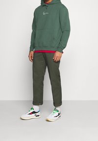Sudadera con capucha verde con cordones y bolsillo delantero, sobre una camiseta roja. Pantalones verde oliva y zapatillas multicolor con base blanca.
