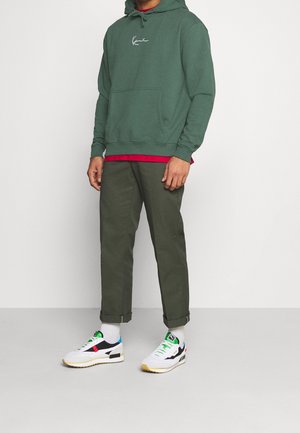 Sudadera con capucha verde con cordones y bolsillo delantero, sobre una camiseta roja. Pantalones verde oliva y zapatillas multicolor con base blanca.