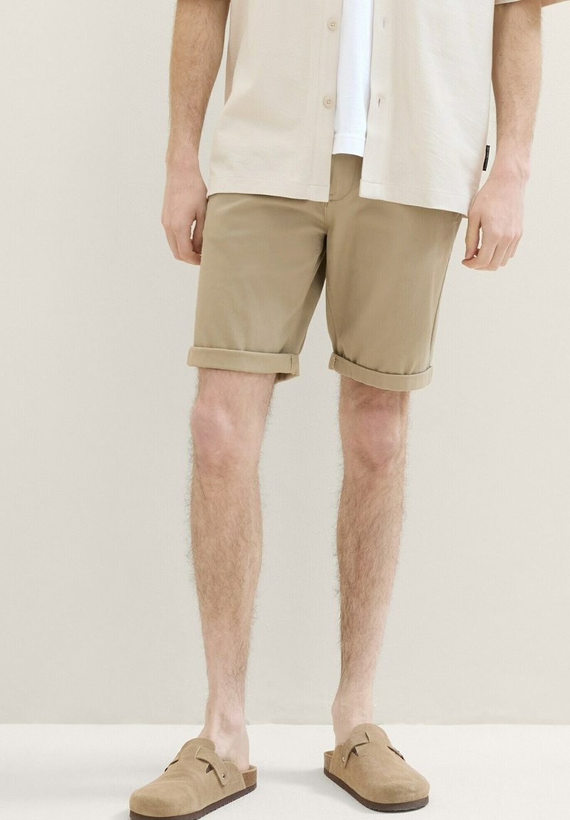 Pantaloni scurți din bumbac bej cu manșete răsucite, asociați cu o cămașă de culoare deschisă și pantofi slip-on din suede în nuanță de bej. Design simplu și casual.