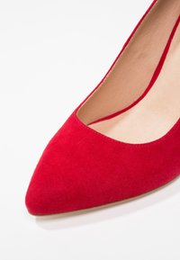 Escarpin en daim rouge avec un bout pointu et un décolleté bas, présentant un intérieur lisse de couleur claire et une silhouette élancée.