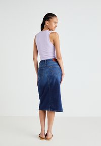 Naine palmikutega juustega, kes kannab lavendlivärvi varrukateta toppi, põlveni ulatuvat sinist teksaseelikut ja pruune sandaale, seisab valge tausta ees.