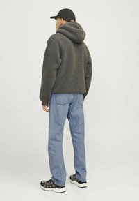 Sudadera de felpa gris con capucha ajustable, combinada con jeans de color azul claro y zapatillas negras con textura. Posición de pie, vista desde la parte de atrás.