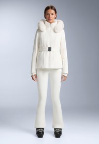 Jachetă albă de schi cu glugă din blană sintetică, textură cu nervuri, fermoar pe față și talie cu curea, purtată cu pantaloni de schi albi evazați și cizme de schi negre.