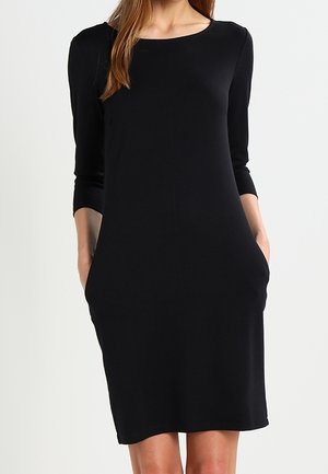 Kobieta ubrana w czarną sukienkę sięgającą do kolan z długimi rękawami i bocznymi kieszeniami, stojąca na tle jasnego, jednolitego tła.