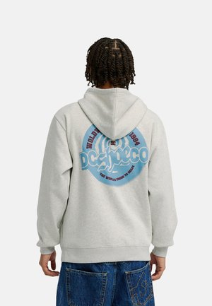 Person with short twists wears a light gray hoodie featuring a large blue and red graphic logo and baggy blue jeans with a back pocket design.
