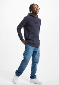 Ung mand med fletninger iført en marineblå hættetrøje, blå jeans og hvide sneakers, stående med det ene ben let bøjet.