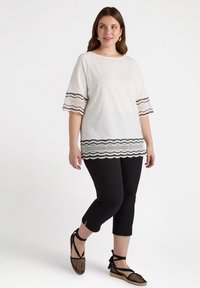 Weißes Oberteil mit schwarzen Spitzenakzenten, mit weit ausgestellten Ärmeln, kombiniert mit schwarzen, knöchellangen Hosen und schwarzen, geflochtenen Espadrilles.