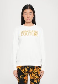 Felpa bianca con "VERSACE JEANS COUTURE" in lettering dorato. Polsini e orlo a coste, abbinata a pantaloni neri e dorati a fantasia.